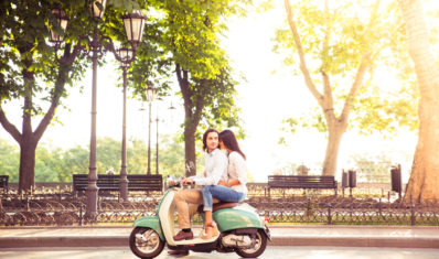 Sideview of a trendy couple riding a scooter in the morning. Sun is shining through trees